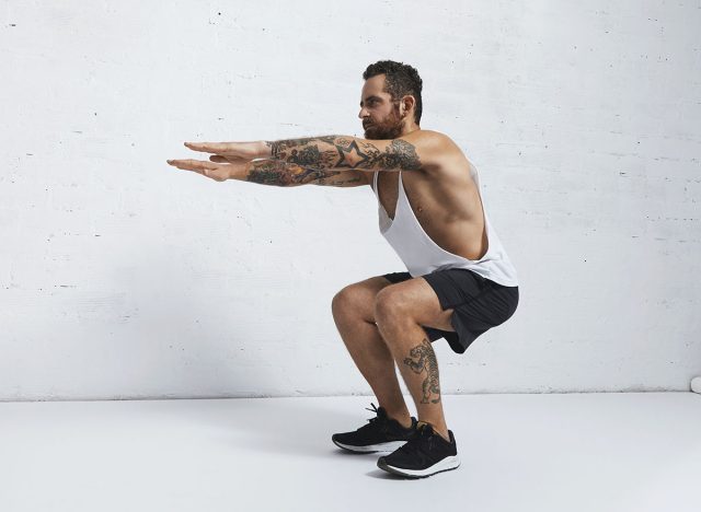 Strong tattooed in white unlabeled tank t-shirt male athlete shows calisthenic moves Squat hold plank exercise
