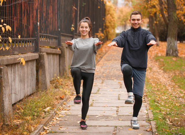 A young sports couple doing exercises standing on one leg and balancing themselves with hands, in an autumn park.