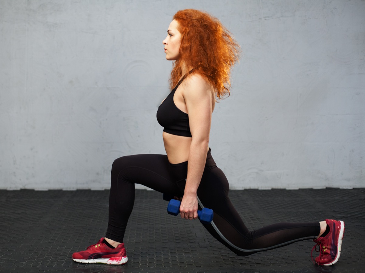 Young redhead woman performing frontal lunge with the dumbbells at the gym