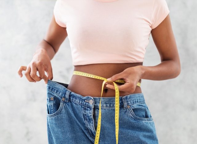 Closeup of black woman in old big jeans measuring her waist with tape, showing results of slimming diet or liposuction, grey background. Young lady promoting healthy nutrition for weight loss