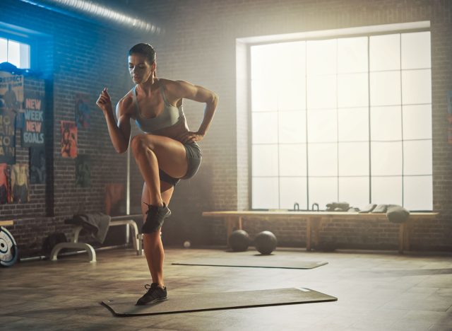 Strong and Fit Beautiful Athletic Woman in Sport Top and Shorts is Doing March and Twist Exercises in a Loft Style Industrial Gym with Motivational Posters. It's Her Fitness Training Workout.