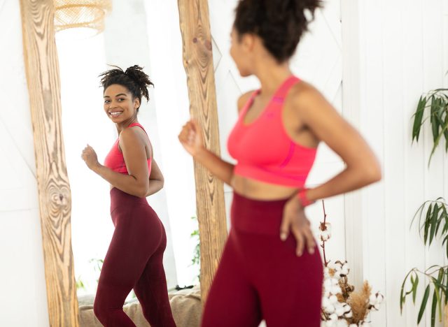 Successful Slimming. Joyful Fit African American Woman After Weight Loss Looking At Reflection In Mirror, Happy About Losing Excess Kilograms Standing At Home. Selective Focus