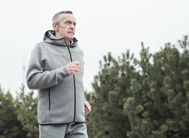 Serious senior Caucasian man in gray sport suit swinging arms while running alone in forest