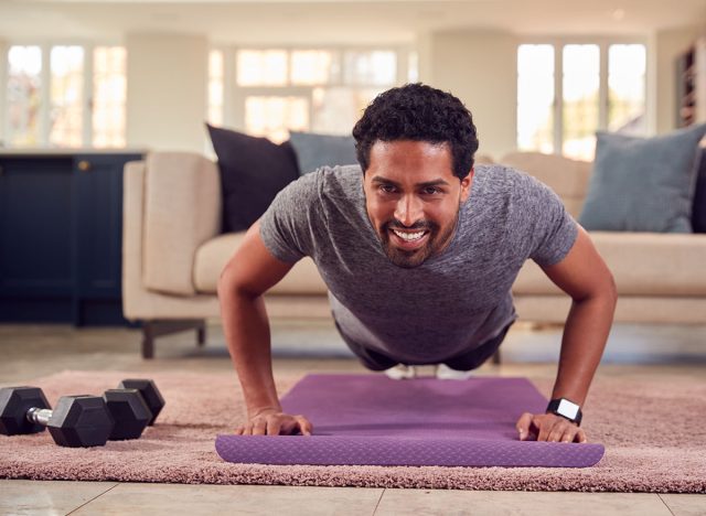 Man In Fitness Clothing At Home In Lounge Doing Press Ups And Exercising With Hand Weights