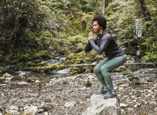 Beautiful mature African American female runner doing squat exercises after jogging near the river in nature.