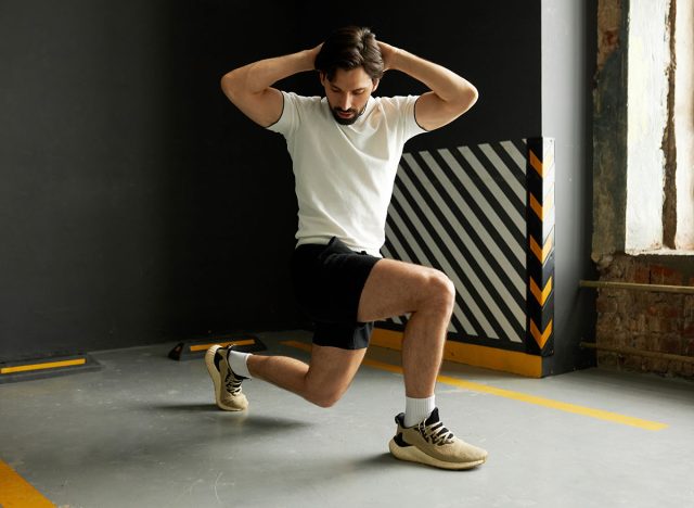 Side view of male athlete in active wear preparing for championship training his legs and full body muscles at gym, lunging forward, putting hands behind head, warming up before hard workout