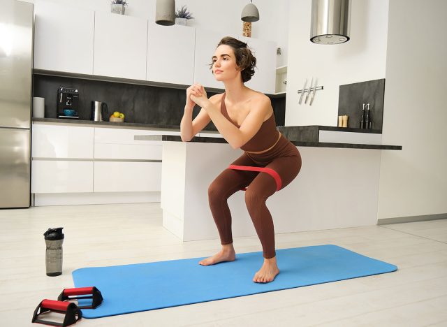 Fitness at home. Young sportswoman, girl in activewear, doing squats in her kitchen, workout indoors on rubber mat, using elastic rubber band for exercises.