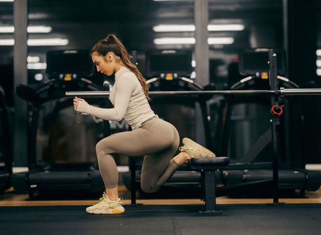A sportswoman in shape is doing split squats on a bench in a gym.