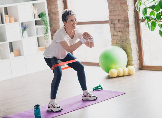 Full length photo of nice pensioner woman resistance band squats sportswear home exercising routine living room healthy lifestyle