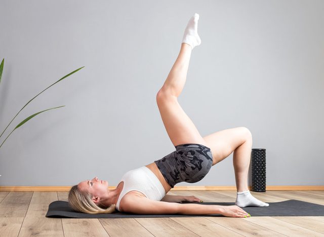 Attractive young fit woman lying on back and does shoulder bridge exercise with one leg up. Pilates black mat at home