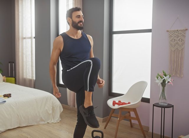 A bearded man in athletic attire exercising in a well-lit, modern bedroom, lifting his knee for a workout routine.