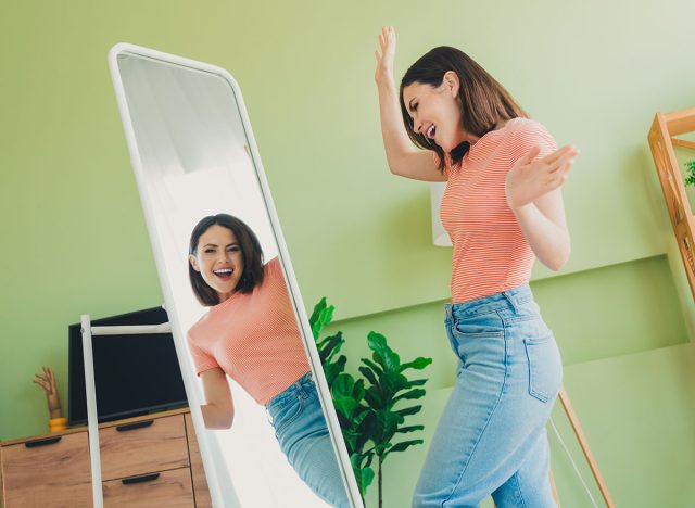 Happy young woman in striped t-shirt enjoying her reflection in a full-length mirror at home, surrounded by plants and modern furniture