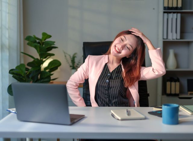 Asian female manager taking a break from work and stretching neck while sitting at desk in modern office