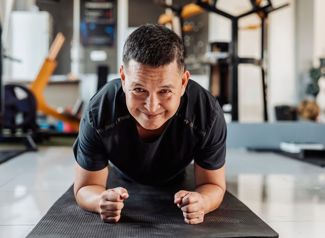 Southeast Asian man in his 50s holds a plank on mat with a confident smile, demonstrating strength and stability in core training.