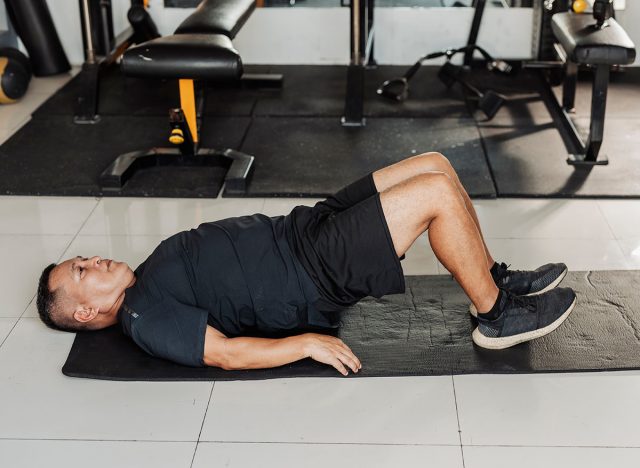 Southeast Asian man in his 50s performs glute bridge exercise on mat to activate glutes and core muscles during workout.