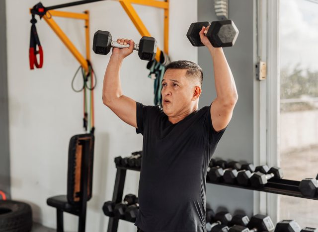 Southeast Asian man in his mid 50s performs an overhead dumbbell shoulder press at an open-air gym with proper form and dedication.