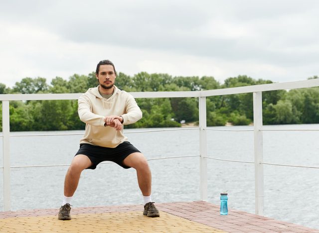 A sporty man in a hoodie performs squats outdoors, embracing nature while working out.