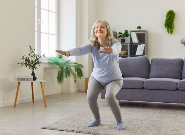 Squat exercise of senior woman training at home, front view. Old happy lady squatting with arms stretching forward and smile, active sport workout in carpet in living room. Rehab, wellness concept
