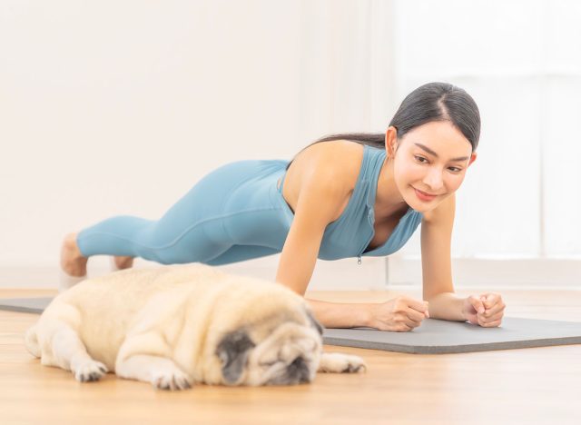 Happy Asian woman in sportswear practicing yoga wellness Asana pose and plank exercise on yoga mat in bright room. Healthy woman enjoy sport training workout relaxation exercise with pet dog at home.