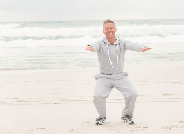 Mature man squatting with arms extended wearing light gray hoodie and sweatpants on sandy shoreline. Fitness, wellness, outdoor, casual, relaxation, health, tranquility