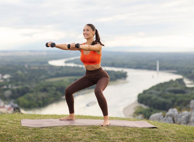Athletic lady doing squat exercises with dumbbells outdoors, having morning training on top of mountain. Active lifestyle concept