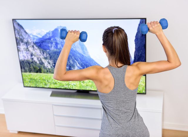 Fitness at home - woman working out in front of tv. Back of a young female adult watching television during her workout, lifting weights to tone arms and shoulders, following an exercise video.
