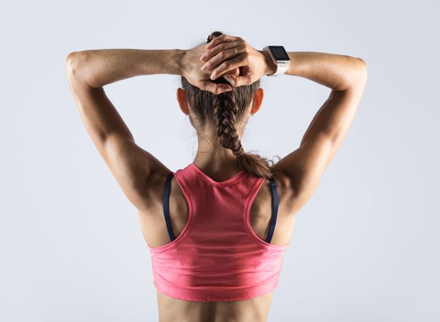 Attractive young fitness person with perfect body wearing red sportswear bra standing against grey background. Well-built sporty model girl wearing smartwatch posing with hands behind head. Back view