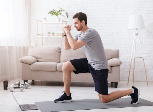 Sport activities at home. Young man watches online exercises and doing lunges in bright living room interior