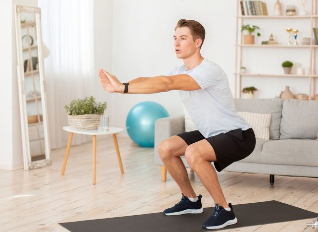 Keeps fit during covid-19 quarantine at home.Young man in sportswear and with fitness tracker squats in interior of living room in morning