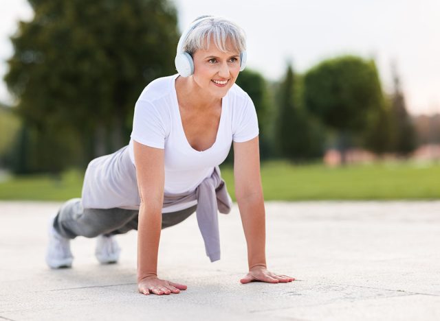 Stay active in retirement. Positive elderly athlete in wireless headphones and casual sportswear training outside in city park, mature active woman doing push ups outdoor in open air