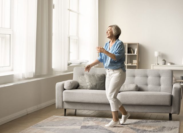 Happy active elderly mature woman dancing in living room alone, enjoying motion, music, entertainment, retirement in modern home interior, having fun. Full length wide shot