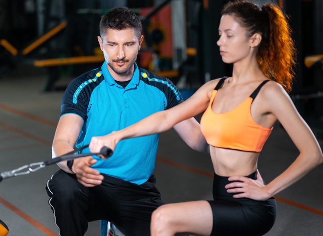 young woman exercising single arm row kneeling with personal trainer man in gym