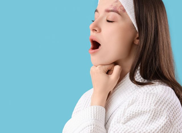 Beautiful woman in bathrobe doing face building exercise on blue background, closeup