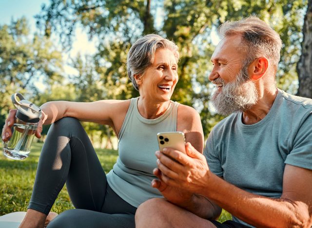 Gadget for recreation after sport. Cheerful couple with grey hair relaxing at park after workout and surfing internet on mobile. Beautiful woman holding water bottle while bearded man using cellphone.