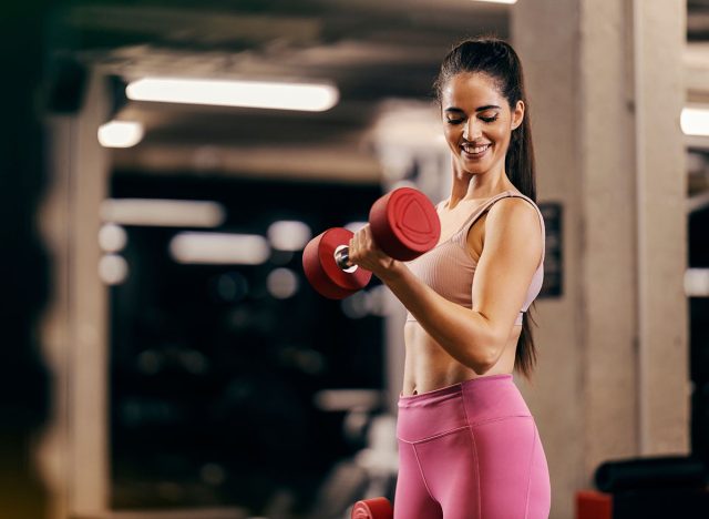 A muscular sportswoman is doing dumbbells curls in a gym and flexing muscles.