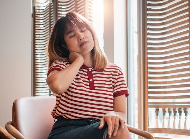 A woman in a striped shirt is sitting in a chair with her head tilted to the side. She is holding her neck and she is in pain. The scene suggests a moment of discomfort or distress for the woman