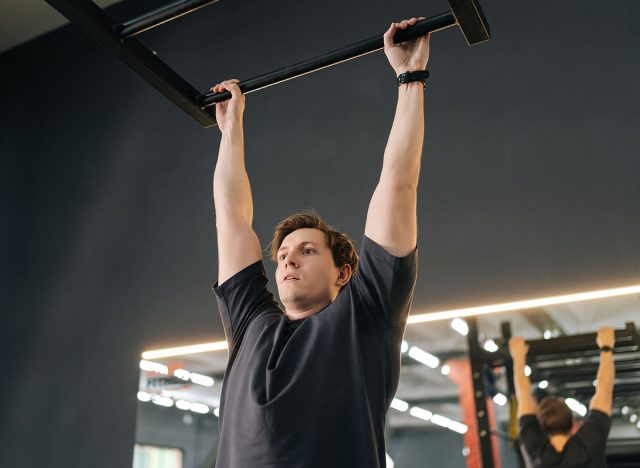 Exhausted muscular male hanging on bar and does one pull-up during sport workout training in modern gym. Tired motivated beginner sportsman showing hard expression on face in pulling up own body.
