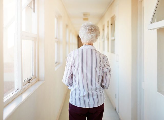 Senior, back and woman walking with corridor in retirement home by window for morning in apartment. Elderly female person or resident having a stroll down hallway, passage or path in flat or building
