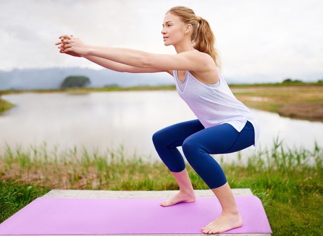 Fitness, woman and workout with squats outdoor for exercise, training and physical activity with balance. Person, stretching and practice on yoga mat at lake for endurance, healthy body and wellness
