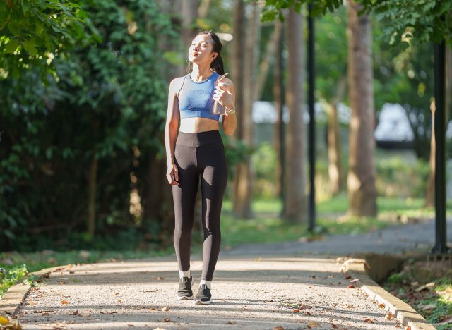 Young asian woman, dressed in sportswear, walks along a park path, holding a water bottle and enjoying the warmth of the sun after her workout