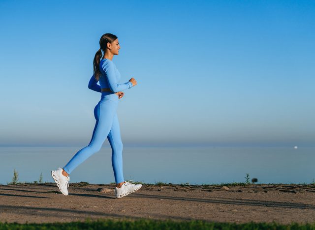 A woman in a vibrant blue outfit briskly walks along a serene waterside path, basking in the calmness of a clear morning sky.