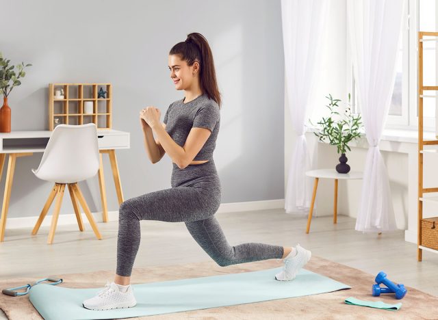Active sporty young woman having fitness workout at home, doing front lunges on yoga mat in living room. Athletic smiling slim girl in sportswear strengthening her body, maintaining healthy routine.