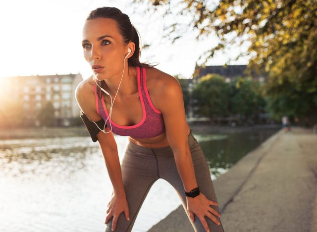 Female runner standing bent over and catching her breath after a running session along lake in city. Young sports woman taking break after a run.