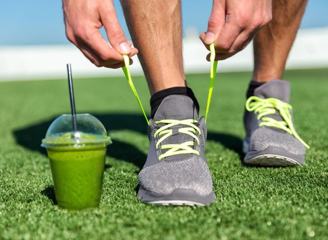 Green smoothie fitness man lacing running shoes, Athlete runner with green vegetable detox juice getting ready for morning run tying running shoe laces on grass. Fitness and healthy lifestyle concept.