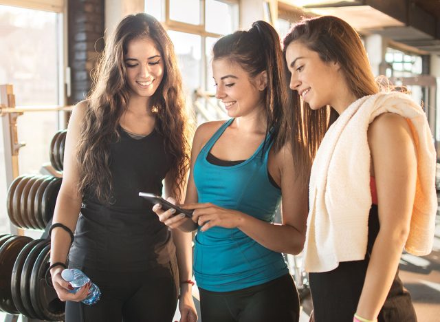 Group of sporty female friends using app on mobile phone, looking at screen of smart phone, texting, watching photos while resting after practice, on break between exercises in gym.