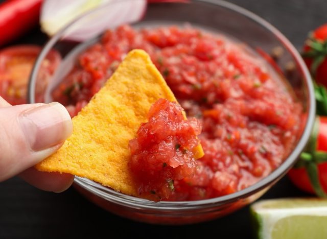 Woman dipping nacho chip into spicy salsa sauce at table, closeup