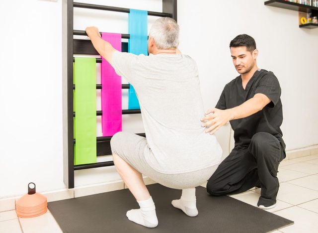 Young physical therapist assisting mature man doing squats in clinic