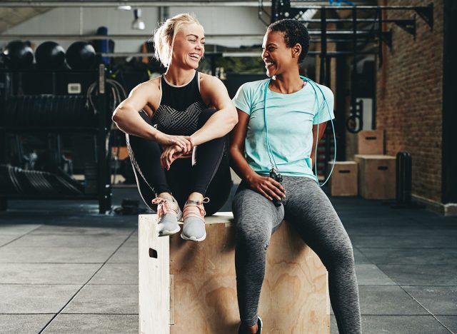 Two fit young female friends in sportswear laughing together while sitting on a box at the gym after a workout session