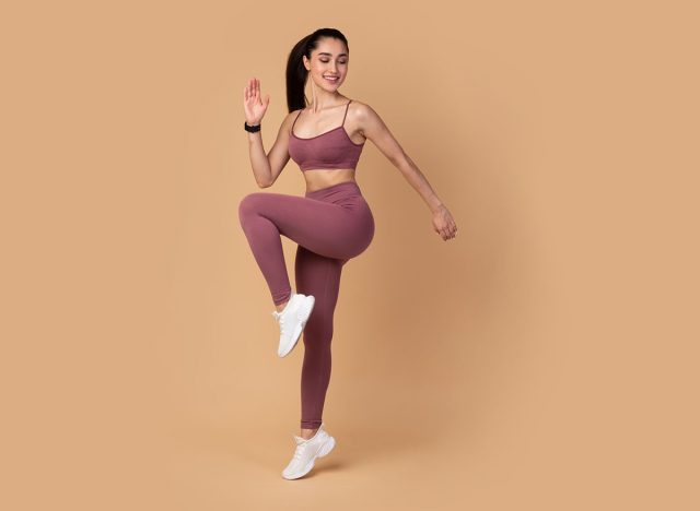 Warmup. Full body length shot of smiling young fitness model in sportswear standing on one leg and touching knee with her elbow, doing high knees exercise isolated over pastel studio background