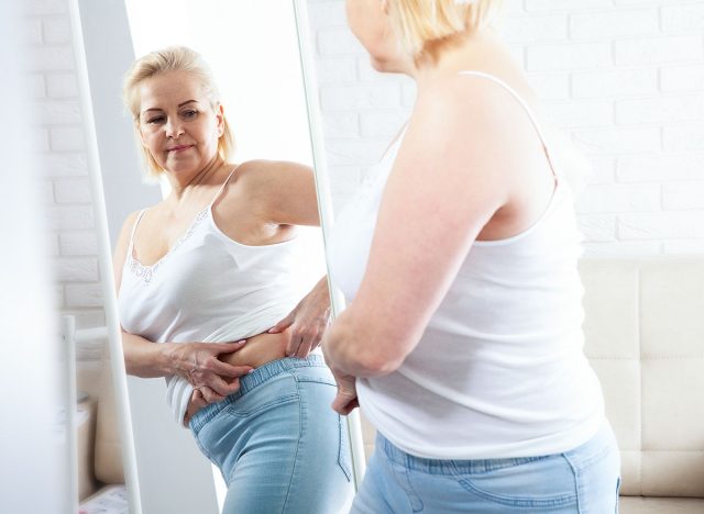 Displeased middle-aged woman looking at herself in the mirror and pinching stomach fat in domestic interior. Sad plus size woman looking her reflection in the mirror with sorrow at home.